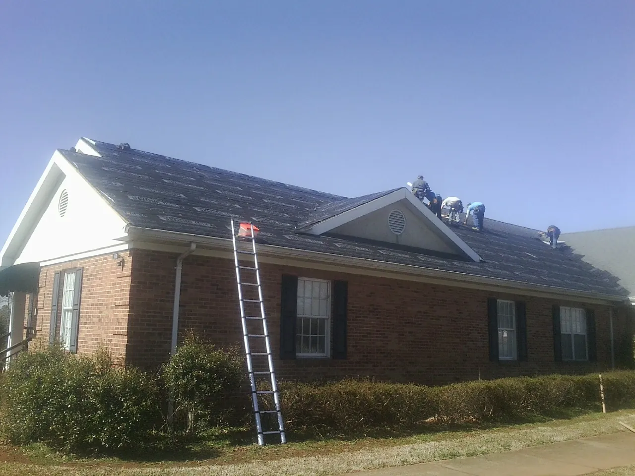 Crew tearing shingles off a brick ranch roof with a ladder set — Emergency Roof Repair work by our crew in Tafton