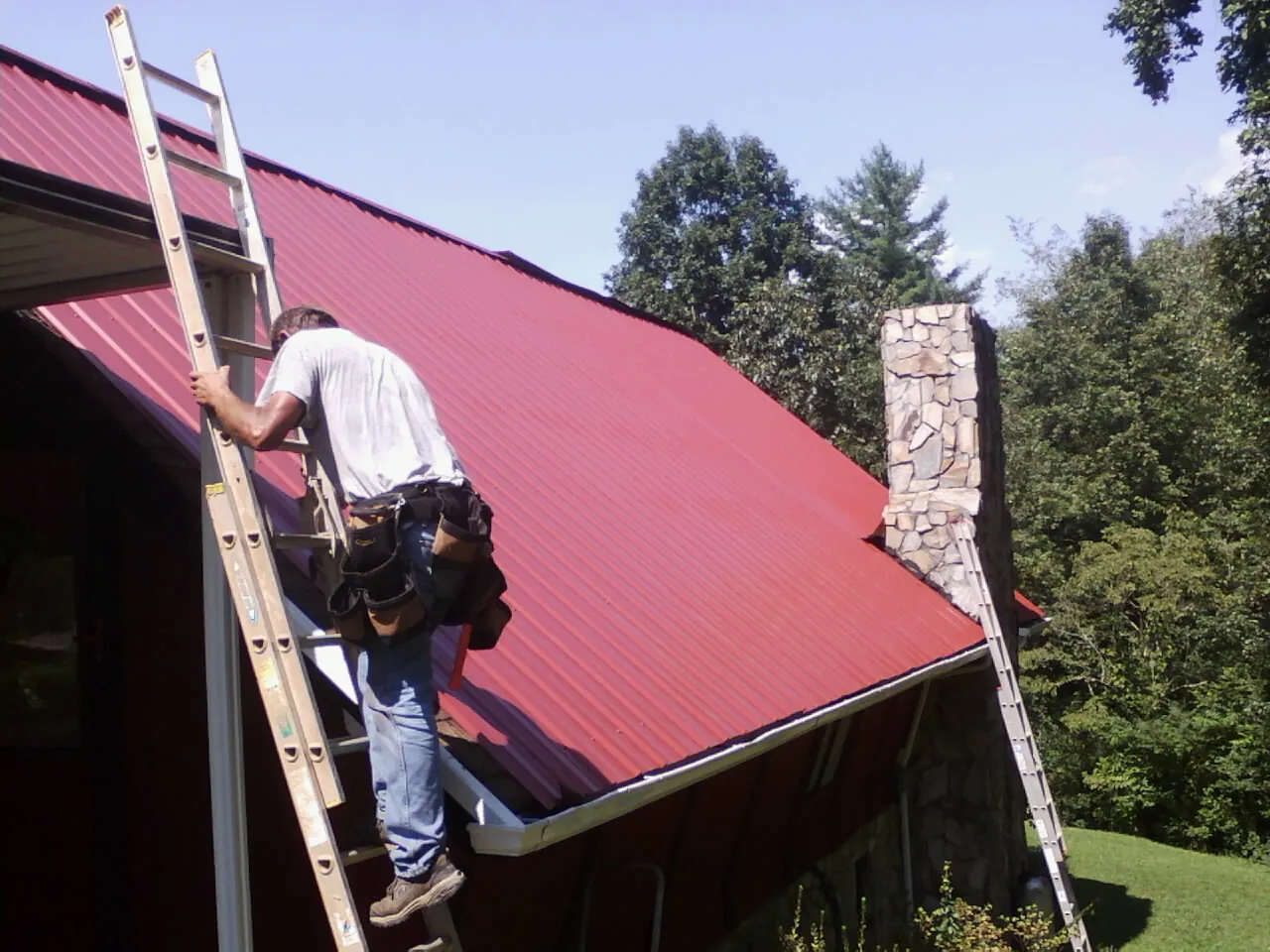 Roofer on a ladder beside a red metal roof and stone chimney — Soffit & Fascia Repair by Roofing Brothers in Tafton